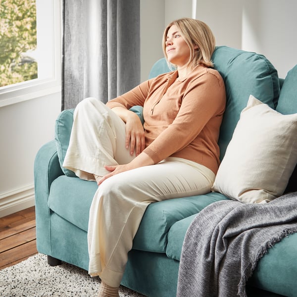 Eine Frau entspannt sich auf einem blaugrünen VINLIDEN-Sofa, das mit einer beigen Decke bezogen ist, in der Nähe eines Fensters mit durchsichtigen Vorhängen.