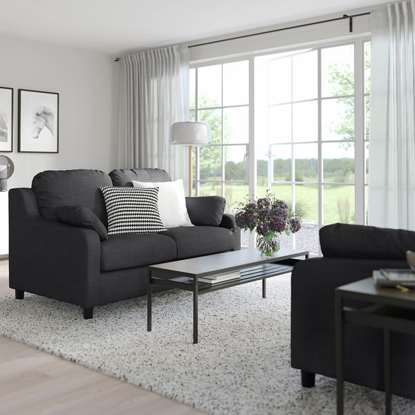 Modernes Wohnzimmer mit grauem VINLIDEN-Sofa, schwarz-weißen Kissen, Couchtisch aus Glas und großem Fenster mit Gartenblick.