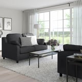 Modernes Wohnzimmer mit grauem VINLIDEN-Sofa, schwarz-weißen Kissen, Couchtisch aus Glas und großem Fenster mit Gartenblick.