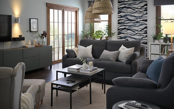 Modernes, gemütliches Wohnzimmer mit grauem VINLIDEN-Sofa, gestreiften Kissen, hölzernem Couchtisch, großen Fenstern und dekorativen Pflanzen.
