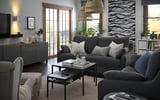 Modernes, gemütliches Wohnzimmer mit grauem VINLIDEN-Sofa, gestreiften Kissen, hölzernem Couchtisch, großen Fenstern und dekorativen Pflanzen.
