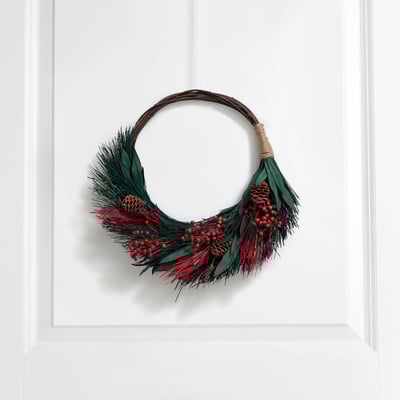 An einer weißen Tür hängt ein Winterkranz mit Grün, roten Beeren und Tannenzapfen, der einen natürlichen, einladenden Charme ausstrahlt.