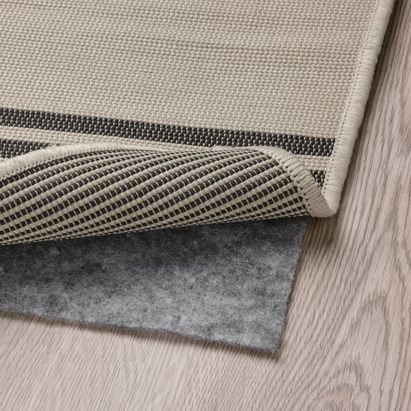 Beige gestreifter Teppich mit schwarzer Rückseite auf Holzboden, dunkelgraue Unterlage für Grip und Komfort, Innen- und Außenbereich.