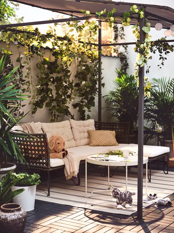 Terrasse mit beigem Teppich, gepolsterten Sitzgelegenheiten, Couchtisch, Pflanzen, Pergola und Beleuchtung.