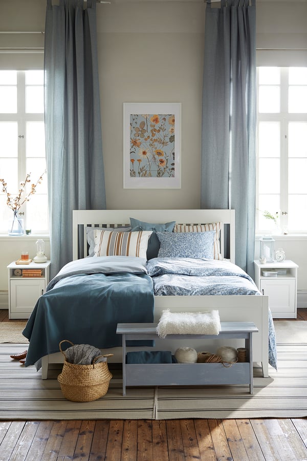 Blaugraues Schlafzimmer mit weißem Bett, Nachttischen, grauem Teppich, blauen Vorhängen, Kunst und Korb zu Fuß.