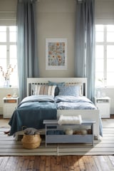 Blaugraues Schlafzimmer mit weißem Bett, Nachttischen, grauem Teppich, blauen Vorhängen, Kunst und Korb zu Fuß.