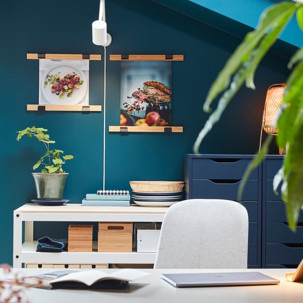 Homeoffice mit grünen Wänden, weißem Schreibtisch, Laptop, vkb-Posteraufhängern mit Küchenfotos und Pflanzen.