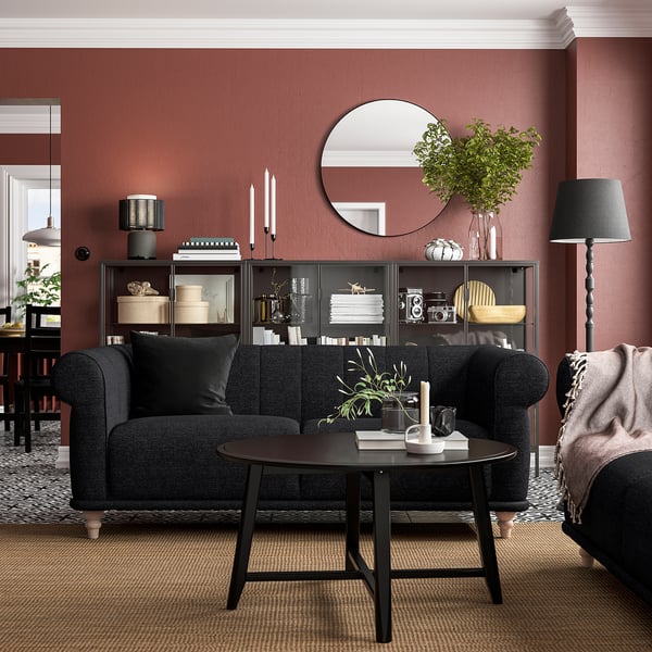 Gemütliches Wohnzimmer mit rostroten Wänden, schwarzem Sofa und deren Dekor, einschließlich Pflanzen und stilvoller Tischlampe.