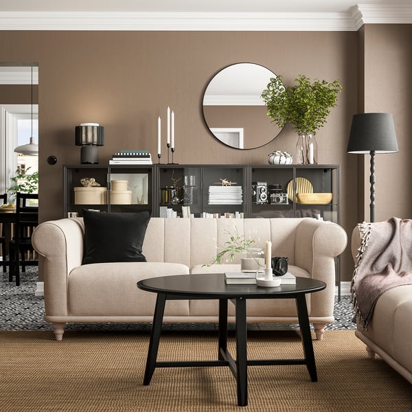 Wohnzimmer mit beigem VISKAFORS-Sofa, schwarzem Couchtisch, rundem Spiegel, Stehlampe und dekorierten Regalen.