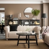 Wohnzimmer mit beigem VISKAFORS-Sofa, schwarzem Couchtisch, rundem Spiegel, Stehlampe und dekorierten Regalen.