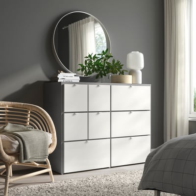 Modernes Schlafzimmer mit VISTHUS Kommode, Spiegel, Lampe, Pflanze, Korbstuhl, grauem Bett, Fenster.