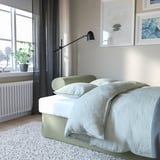 Gemütliches Schlafzimmer mit grünem VRETSTORP-Schlafsofa, moderner Lampe und Wandkunst. Ein Heizkörper und eine Pflanze am Fenster.