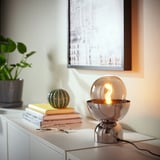Nickel-plated ACKJA table lamp with smoke glass on white furniture, books, plant, and frame.