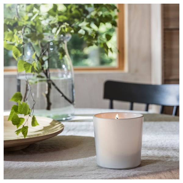 A lit ADLAD scented candle, white ceramic, on a textured tablecloth, next to a vase with green leaves, and wooden chair.