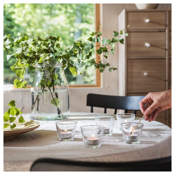 Hand lighting small glass candles on table with flowers and wooden furniture.
