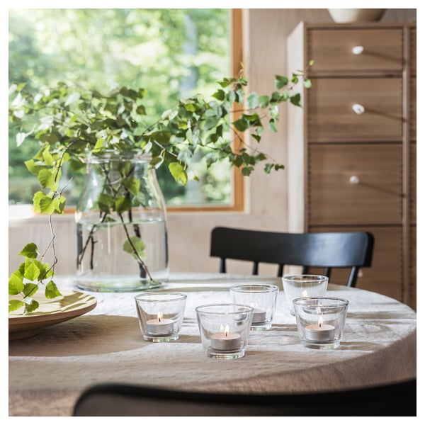 Cozy table setting with four lit glass candles, green plant, wooden table, and black chair.