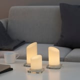 Three lit candles of varying heights on glass bases sit on a table in a living room setting, creating a warm ambiance.