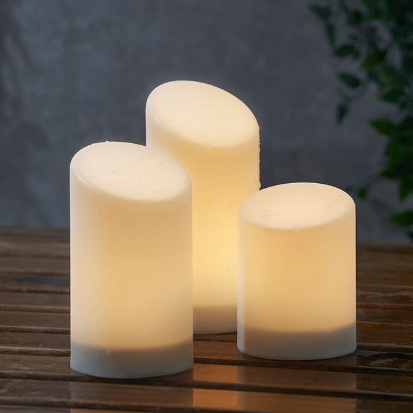 Three white candles of varying heights on a wooden surface. They emit a warm, flickering light, safely simulating real candles.
