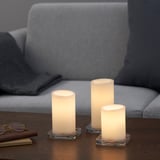 Three led candles of varying heights sit on a wooden table in front of a grey couch, one flickers warmth.