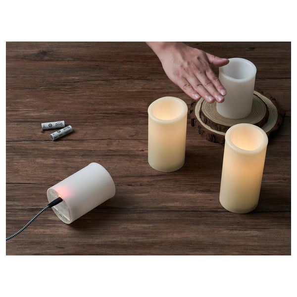 Image battery-operated led candles on a wooden surface. One candle is lit, another off. Batteries lie nearby. A hand adjusts the unlit candle.