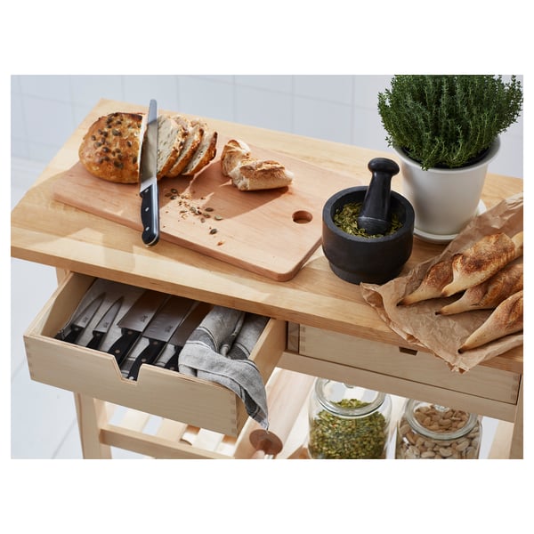 Wooden kitchen cart with open drawer showing knives, loaf of bread, mortar with pestle, plant, and jars with spices.
