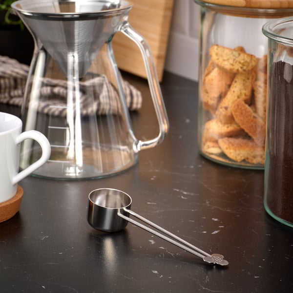 A kitchen counter shows a glass pour over coffee maker, a white mug on a coaster, a metal coffee scoop, and jars filled with biscuits and tea bags.