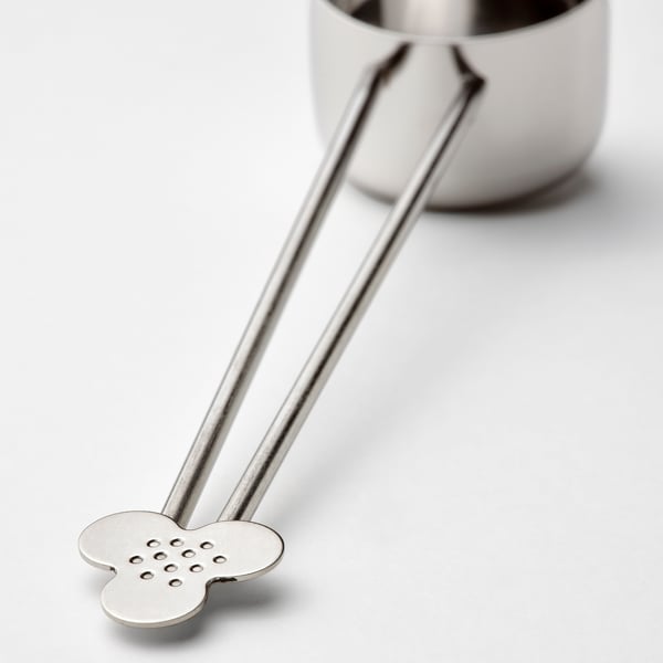 Silver tea infuser with long handles and flower-shaped perforated tip, resting near a mug.