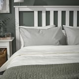 Bed with white ÄNGSLILJA pillows, grey cover, against greenish wall. Nightstand beside it with decor.