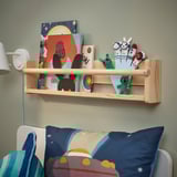 Wooden shelf AFTONSPARV holds colorful books and playful figurines above a bed with blue pillows.