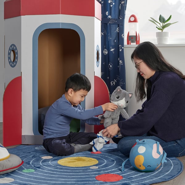 A child plays with a woman, both holding a plush grey cat in a colorful, space-themed room.