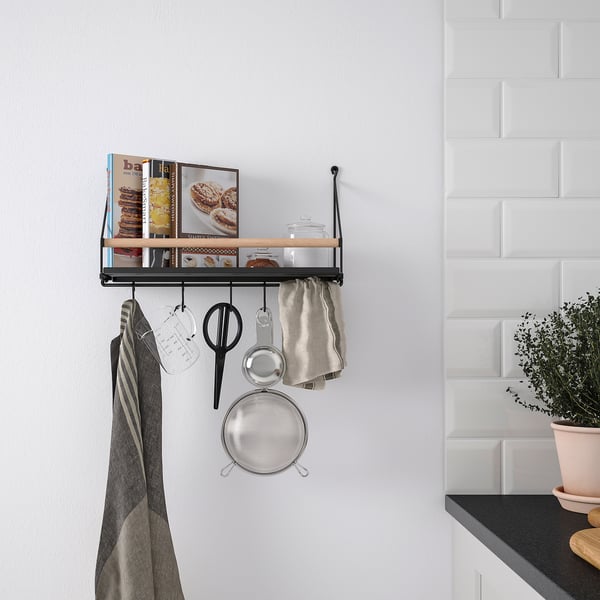 Black metal wall organizer with hooks and wooden shelf, holding kitchen items and cookbooks, beside a counter with a plant.