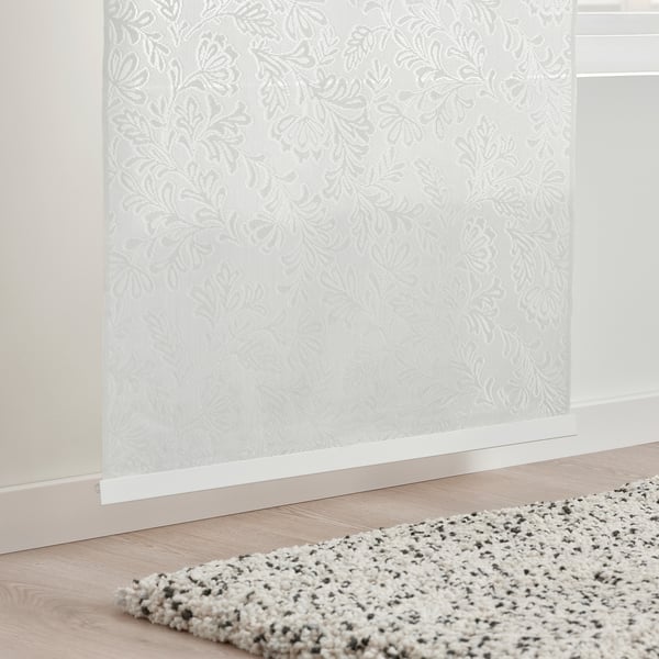 Sheer curtain with leaf pattern allows filtered light. Positioned near a speckled rug on a wooden floor.