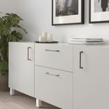 White cabinet with silver ANDERSFORS handles, sleek and minimalist design. Its larger than the potted plant and smaller than the stacked books.