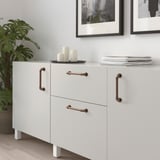 Modular white cabinet with ANDERSFORS copper handles, slim design, fit neatly against wall, topped with books and candles, beside large plant.