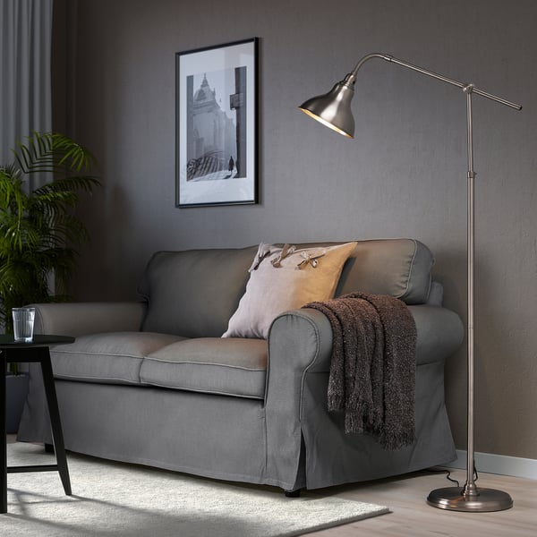 A tall ANKARSPEL lamp in a cozy living room beside a grey couch with pillows and a blanket. The brushed pewter finish complements the decor.