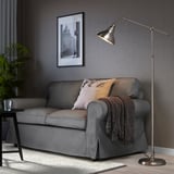A tall ANKARSPEL lamp in a cozy living room beside a grey couch with pillows and a blanket. The brushed pewter finish complements the decor.