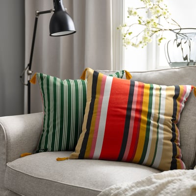 Gray sofa with multi-stripe AROMATISK cushion near lamp and window, flowers in background.