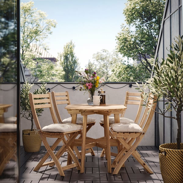 Small outdoor dining set with a brown table and chairs, cushions on chairs. Surrounded by plants and flowers.