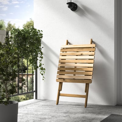 A wooden folding chair leans against a white wall outdoors. A plant hangs nearby, adding greenery to the scene.