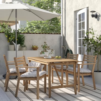 Outdoor dining set with wooden table and chairs. Chairs have white cushions. Set is on a patio with a large umbrella. Table has a vase with flowers.