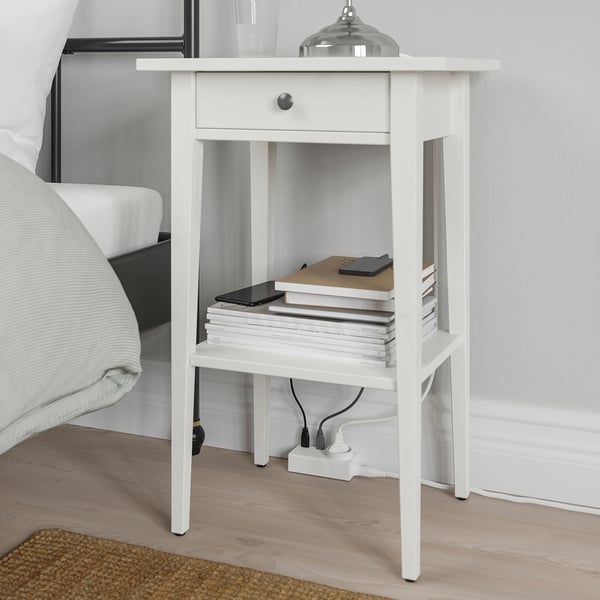 A white nightstand beside a bed with a drawer and an open shelf holding a stack of books and a tablet. A cable management setup is visible below.
