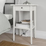 A white nightstand beside a bed with a drawer and an open shelf holding a stack of books and a tablet. A cable management setup is visible below.
