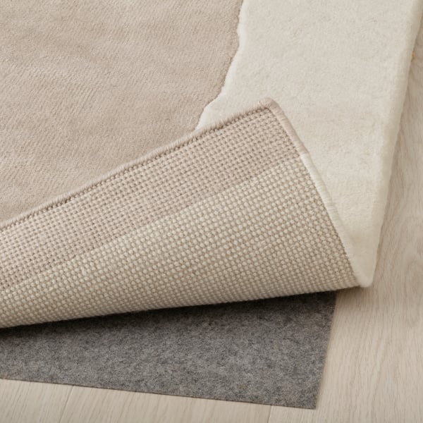 Image shows corner of thick, beige, handmade wool rug with visible texture detail.