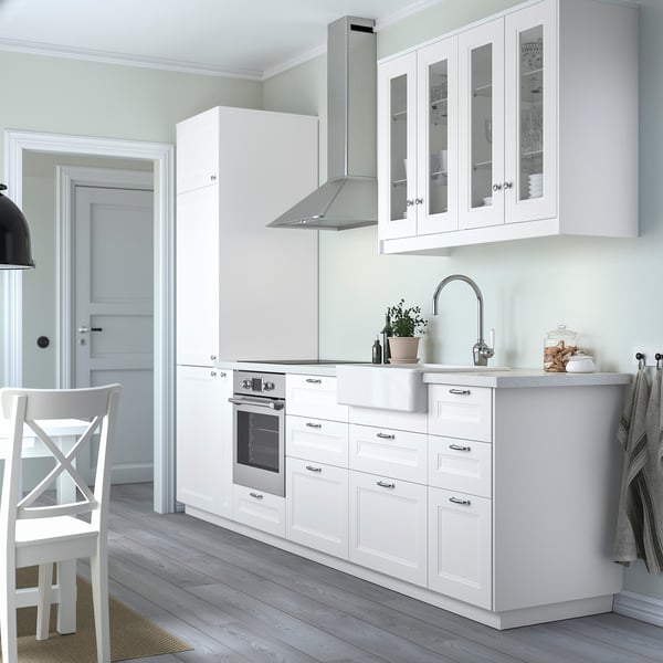 Modern white kitchen with AXSTAD cabinets, stainless fixtures, and glass doors, surrounded by light wood floors and a dining chair.
