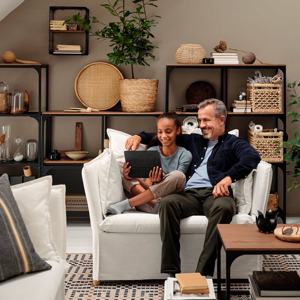 A man and child sit on a white BACKSÄLEN armchair, smiling while using a tablet. Behind them, shelves hold books and plants.