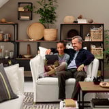 A man and child sit on a white BACKSÄLEN armchair, smiling while using a tablet. Behind them, shelves hold books and plants.