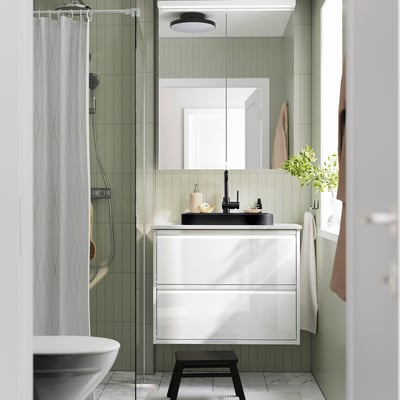 Modern bathroom with semi-recessed black marble basin, white cabinet, green tiles, glass shower, towel hooks, and plant.