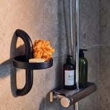 Black BÄSINGEN shower holder with orange sponge and soap bottles, modern design with dark grey finish, mounted on grey tiled wall.