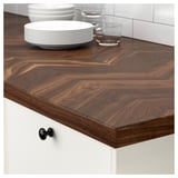 Wooden kitchen counter in dark brown with stackable dishes and glasses. Chevron pattern, sleek design, white cabinets with black knobs.