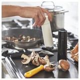 Person seasoning pan of mushrooms with white grinder, black grinder beside, and chopped mushrooms on tray.